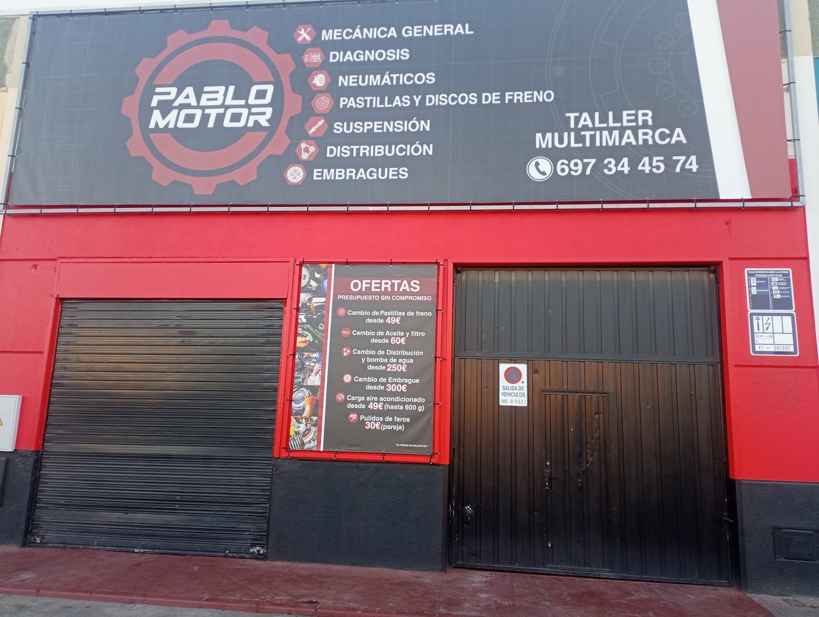 Fachada del taller con las persianas bajadas, el rótulo de servicios y un cartel de ofertas