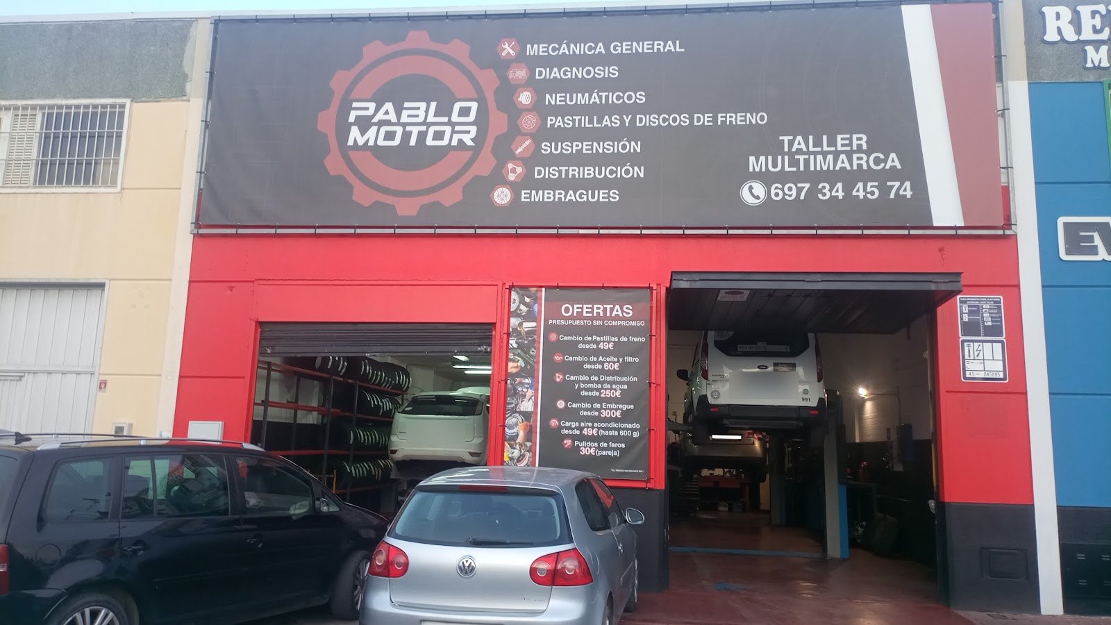 Fachada roja del taller Pablo Motor con su rótulo negro de servicios y el teléfono, y un coche elevado en el foso
